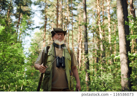 Medium shot of senior Caucasian man with beard on face wearing hat walking in forest with use of wooden stick, copy space 116586073