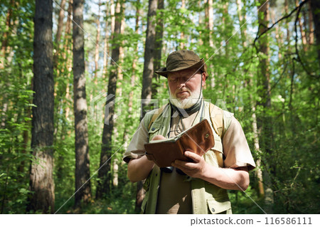 Medium shot of senior Caucasian forester holding notebook writing notes while working outdoors on summer day 116586111