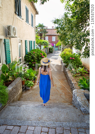 A woman in a blue dress and straw hat walks down a quaint alley lined with green plants and flowers. 116587020