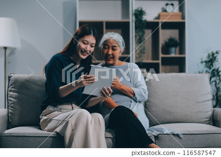 Happy grownup daughter showing content on tablet to mature mother. Two family generations women with digital computer gadget resting on couch together, watching videos, making video call 116587547