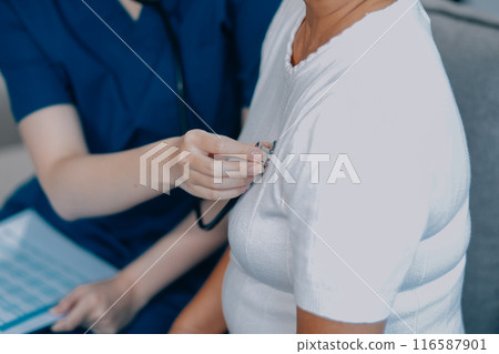 Caucasian female doctor uses stethoscope to check heart rate on elderly Asian patient on sofa. 116587901