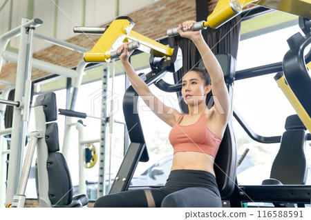 Young woman using gym machine to work on her arms strength during her morning workout at a local gym Young woman using gym machine to work on her arms strength during her morning workout at a local gym 116588591
