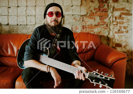 Mature Caucasian rock musician wearing stylish red sunglasses sitting relaxed on leather couch holding guitar and looking at camera, copy space Mature Caucasian rock musician wearing stylish red sunglasses sitting relaxed on leather couch holding guitar and looking at camera, copy space 116588677