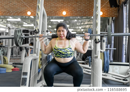 Young overweight woman lifting weight using gym barbell during her morning workout to lose weight at local gym Young overweight woman lifting weight using gym barbell during her morning workout to lose weight at local gym 116588695