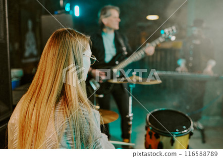 Rear view of unrecognizable woman with long hair playing drums on stage with rock musicians 116588789