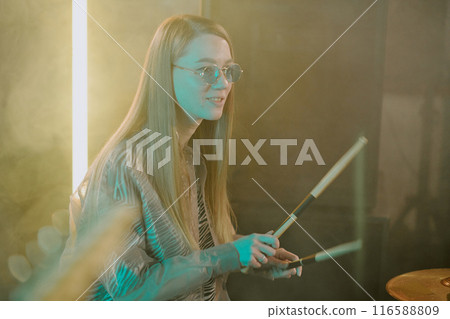 Medium shot of young Caucasian woman with long blond hair wearing stylish sunglasses playing drums in rock band 116588809