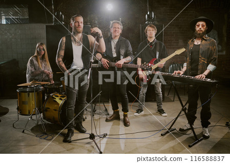 Group of rock male and female musicians playing in rock band posing for camera on stage in modern nightclub 116588837