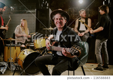 Selective focus portrait of joyful mature Caucasian bassist sitting on chair holding guitar smiling at camera during rehearsal with his band Selective focus portrait of joyful mature Caucasian bassist sitting on chair holding guitar smiling at camera during rehearsal with his band 116588883
