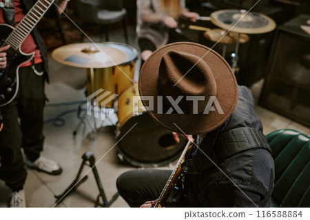 High angle view of unrecognizable man in hat sitting on chair and playing guitar during rehearsal or performance with band High angle view of unrecognizable man in hat sitting on chair and playing guitar during rehearsal or performance with band 116588884