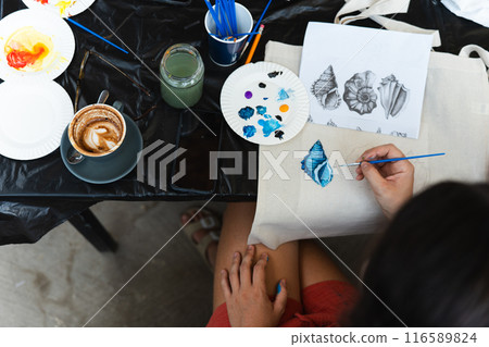 Top view of a woman brush painting a blue shell on a handmade bag with a cappuccino next to it 116589824