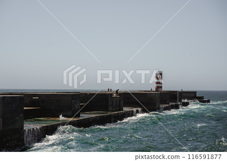 A pier stands in the ocean with a lighthouse in the background A pier stands in the ocean with a lighthouse in the background 116591877
