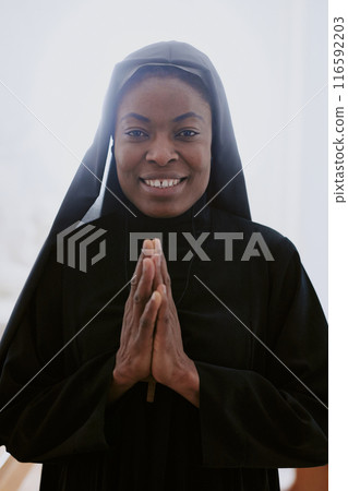 Vertical waist up portrait of happy African American Catholic nun posing for camera with praying hands 116592203