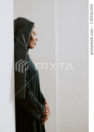 Vertical minimalistic shot of young African American Catholic nun standing still by white wall, copy space Vertical minimalistic shot of young African American Catholic nun standing still by white wall, copy space 116592204