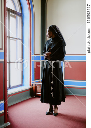 Vertical long shot of young African American Catholic nun wearing black religious clothes standing indoors looking out of window Vertical long shot of young African American Catholic nun wearing black religious clothes standing indoors looking out of window 116592217