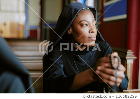 Young African American Catholic nun holding rosary beads sitting on pew and looking away, medium closeup shot Young African American Catholic nun holding rosary beads sitting on pew and looking away, medium closeup shot 116592259