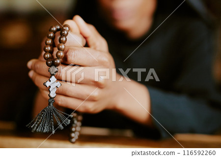 Closeup of hands of unrecognizable young Black nun holding rosary in hands praying to Lord while sitting on pew in church 116592260