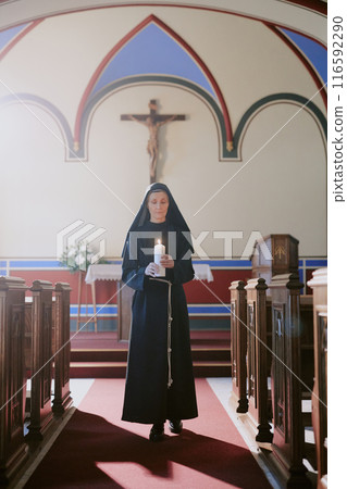 Vertical wide shot of senior Caucasian nun in black habit holding burning candle walking along nave in Catholic church Vertical wide shot of senior Caucasian nun in black habit holding burning candle walking along nave in Catholic church 116592290