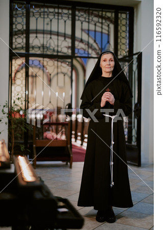 Vertical long shot of mature Caucasian Catholic nun wearing black habit holding cross looking at camera while standing at church narthex Vertical long shot of mature Caucasian Catholic nun wearing black habit holding cross looking at camera while standing at church narthex 116592320