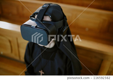 High angle medium closeup of unrecognizable Catholic nun wearing VR headset sitting on wooden pew in church High angle medium closeup of unrecognizable Catholic nun wearing VR headset sitting on wooden pew in church 116592382