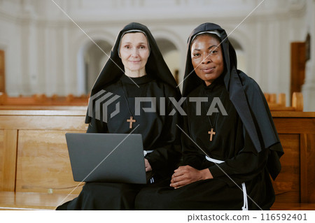 Medium shot of two ethnically diverse nuns sitting on wooden pew in Catholic church, looking at camera while using laptop 116592401
