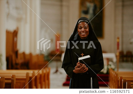 Medium shot of cheerful African American Catholic nun holding Bible book standing at nave, smiling at camera 116592571