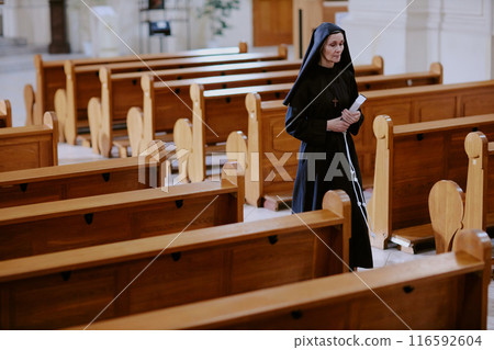 Wide shot of senior Caucasian nun holding prayer book walking along nave in Catholic church, copy space 116592604
