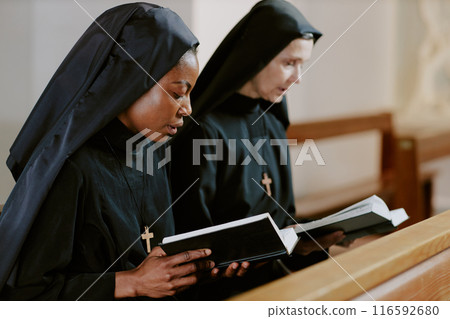 Two ethnically diverse Catholic nuns sitting on pew holding books and saying prayers, medium closeup shot 116592680