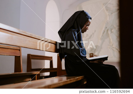 Side view shot of young African American Catholic nun sitting on wooden pew in Catholic church and reading religious literature, copy space 116592694