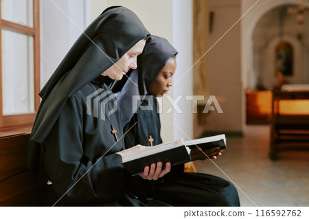 Medium shot of young and mature biracial nuns sitting on bench in church reading religious texts, copy space 116592762