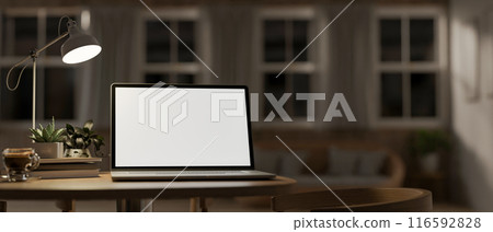A laptop computer mockup, illuminated by light from a table lamp on a wooden table. 116592828