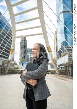 A successful mature Asian businesswoman in a suit is looking away at the sky, standing in the city. 116592904