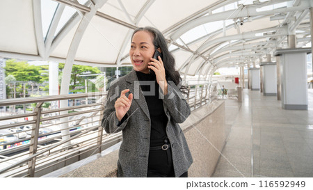 A confident mature Asian businesswoman is talking on the phone while standing on a skywalk. 116592949