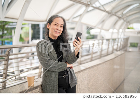An attractive mature Asian businesswoman stands on a skywalk with a smartphone in her hand. An attractive mature Asian businesswoman stands on a skywalk with a smartphone in her hand. 116592956