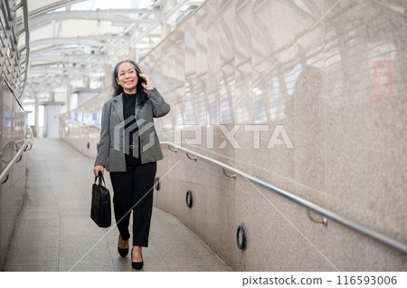 A mature Asian businesswoman is talking on the phone while walking on a skytrain station in the city A mature Asian businesswoman is talking on the phone while walking on a skytrain station in the city 116593006