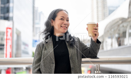 An attractive mature Asian businesswoman with her takeaway coffee cup is standing on a skywalk. 116593021