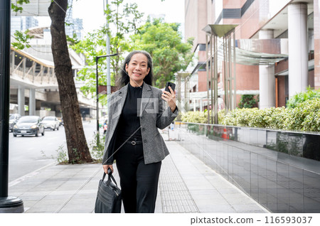 A confident, positive mature Asian businesswoman is walking on a sidewalk in the city. 116593037
