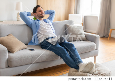 A middle-aged man is seen relaxing on a comfortable couch, engrossed in his laptop screen. He appears calm and focused as he works or browses the internet in a cozy indoor setting. 116593811
