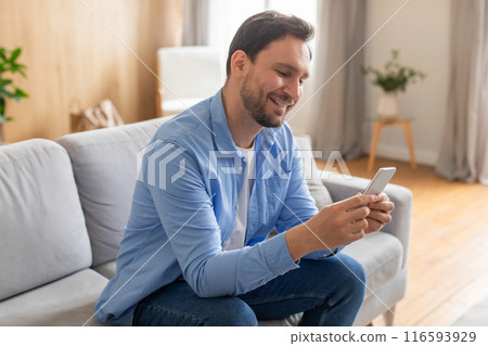 A man is seated on a couch, fixated on his phone screen. He appears absorbed in the digital content displayed on his device. 116593929