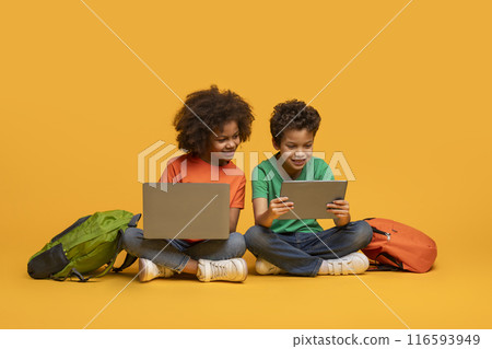 Two young African American children absorbed in a laptop and tablet. Their backpacks rest beside them, hinting at a break from school activities or a moment of leisure after classes. Two young African American children absorbed in a laptop and tablet. Their backpacks rest beside them, hinting at a break from school activities or a moment of leisure after classes. 116593949