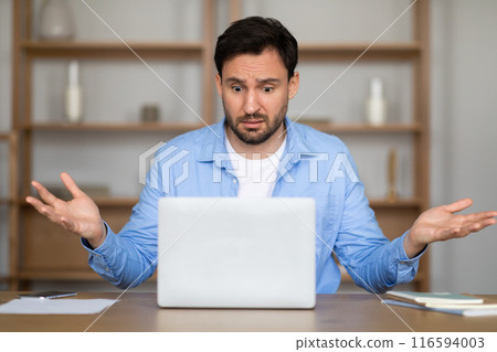 A man in a casual blue shirt sits at a desk, showing a bewildered expression with arms raised in uncertainty as he looks at his laptop screen, likely dealing with a perplexing work situation 116594003