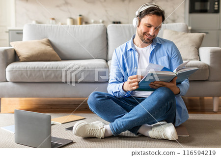 A man is seated on the floor, engrossed in reading a book while simultaneously listening to music through headphones. He appears focused and relaxed in his dual activities. 116594219