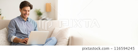 Happy young man sitting on couch at home, using computer laptop, websurfing or working online, panorama with copy space 116594245