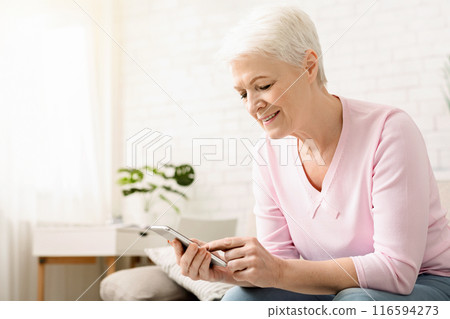 Elderly woman is seated on a couch indoors, engrossed in her cell phone. She appears focused as she interacts with the device, possibly messaging or scrolling through content. Elderly woman is seated on a couch indoors, engrossed in her cell phone. She appears focused as she interacts with the device, possibly messaging or scrolling through content. 116594273