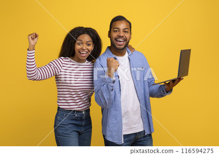 Black couple standing side by side against a yellow backdrop, both exuding excitement and joy. The man holds a laptop in one hand while they both raise their other fists in a triumphant gesture. 116594275