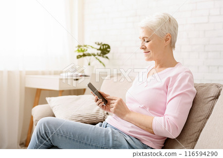 A cheerful senior woman with short hair is sitting comfortably on a beige sofa, holding and looking at a smartphone with a content expression 116594290