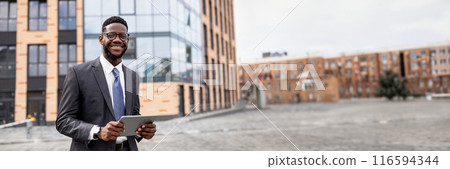 Business news. Happy black businessman using digital tablet walking outdoors near modern office building in urban area cityscape, smiling to camera, copy space, web-banner 116594344