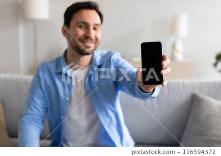 A cheerful man in a blue shirt is extending his arm to proudly display a smartphone with a blank screen, sitting on a sofa in a well-lit, cozy living room environment during the day. 116594372