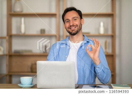 A cheerful man sits in a casual office setting, giving an affirmative okay sign with his hand while looking directly at the viewer. He is wearing a blue shirt and is seated in front of a laptop A cheerful man sits in a casual office setting, giving an affirmative okay sign with his hand while looking directly at the viewer. He is wearing a blue shirt and is seated in front of a laptop 116594393