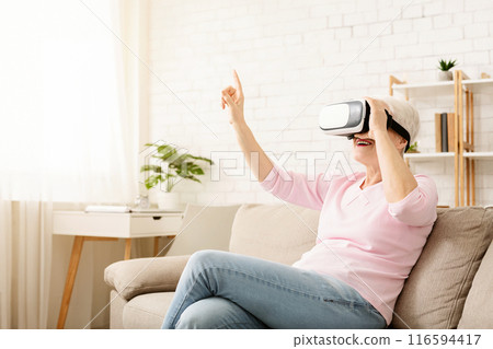 Elderly woman is seated on a couch, wearing a virtual reality headset, engaged in the virtual world, with a hand reaching out in front of her. The room is dimly lit, with the headset glowing blue. 116594417