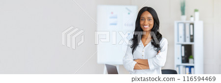 Portrait of happy African American businesswoman standing with crossed arms in front of her desk at modern office. Confident female entrepreneur looking at camera and smiling, copy space 116594469